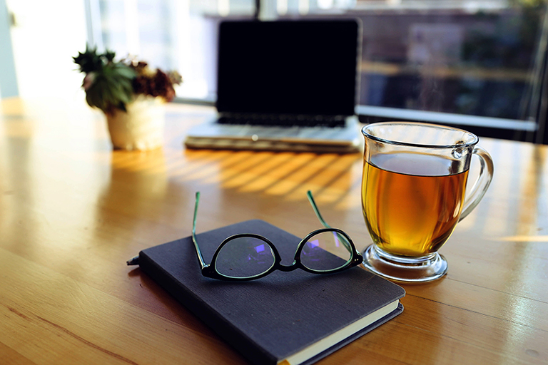 Eyeglasses on top of a black notebook beside a clear glass mug.